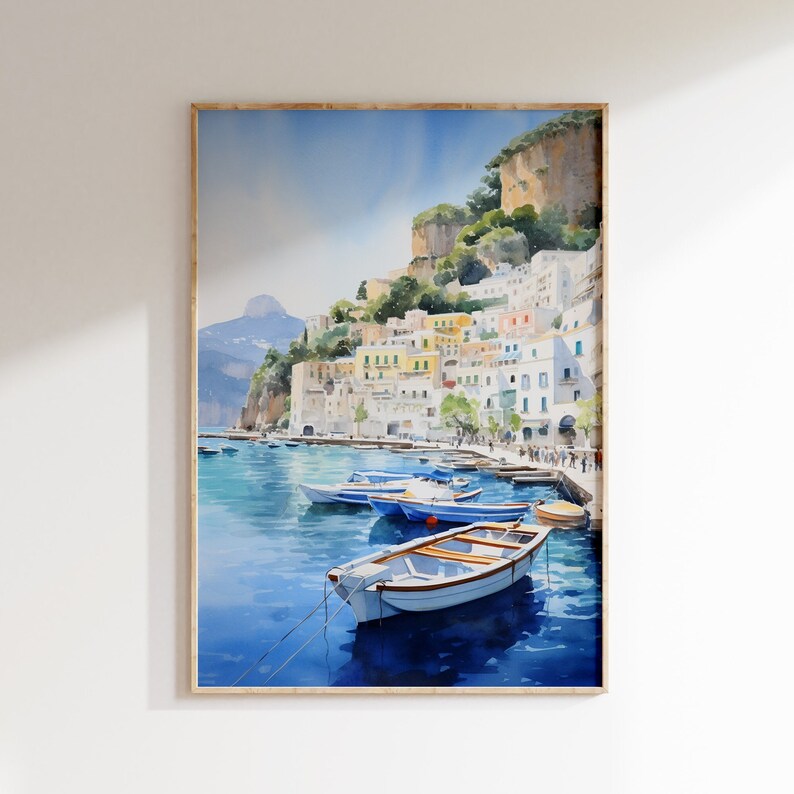 May include: Watercolor painting of a colorful Italian village on a cliffside overlooking a blue bay with several small boats docked. The painting is framed in a light wood frame.