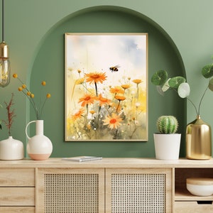 May include: Watercolor painting of orange daisies and a bee in a field. The artwork is framed in a light wood frame and displayed on a wooden cabinet. The background is a green wall with decorative vases and plants.