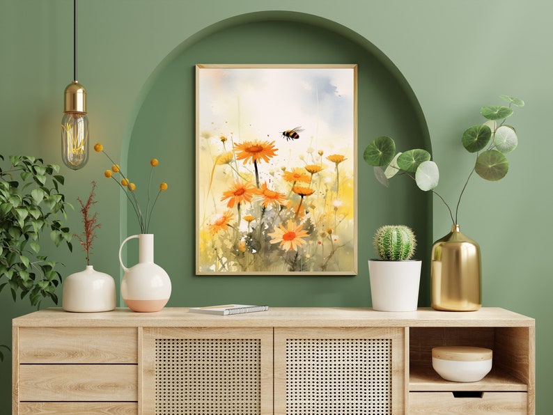 May include: Watercolor painting of orange daisies and a bee in a field. The artwork is framed in a light wood frame and displayed on a wooden cabinet. The background is a green wall with decorative vases and plants.