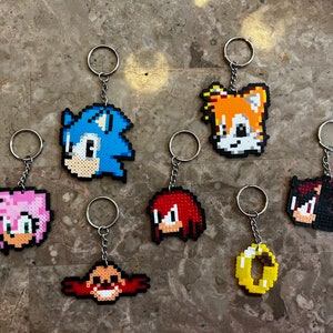 May include: Seven colorful keychains featuring characters from the Sonic the Hedgehog video game series. The keychains are made of plastic beads and feature Sonic, Tails, Knuckles, Amy, Shadow, and a gold ring.