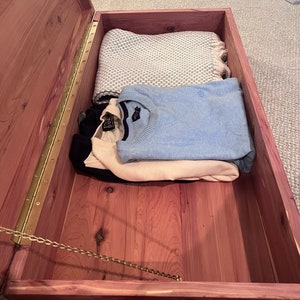 Puede incluir: Un cofre de madera de cedro con un cierre de cadena de latón y una tapa con bisagras. El cofre está abierto y contiene una manta doblada, un suéter azul y un suéter blanco.