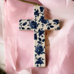 May include: A ceramic cross with a blue and white floral pattern, suspended by a gold cord. The cross is set against a pink background. The floral design features detailed blue flowers and leaves.