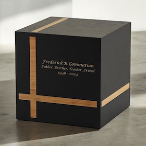 May include: Black wooden cremation urn with a cross design made of light wood. The urn is engraved with the name "Frederick R. Gommarian", the titles "Father, Brother, Teacher, Friend", and the dates "1948 - 2024".