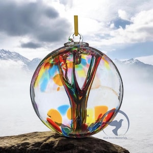 May include: A clear glass memorial orb with a colorful tree design inside. The orb features a vibrant mix of red, yellow, blue, and green colors. A yellow ribbon is attached for hanging. The orb is sitting on a rock with a mountain backdrop.