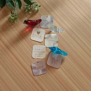 May include: A collection of glass bird figurines in red, clear, and blue, alongside several square cards with printed text and watercolor designs. The cards feature phrases like "One Day at a Time" and "In another time..."