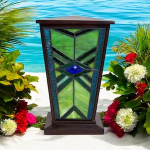 May include: A brown wooden lantern with a stained glass panel featuring green and blue geometric designs. The lantern is set on a square base and has a triangular top. The background includes tropical plants and a view of the ocean.
