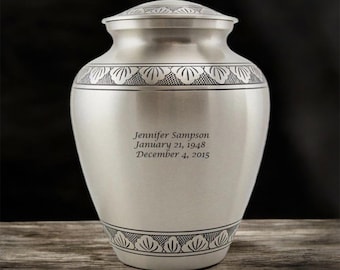 May include: A silver urn with a decorative floral pattern around the top and bottom. The urn has a lid and is engraved with the name "Jennifer Sampson" and dates "January 21, 1948 - December 4, 2015".