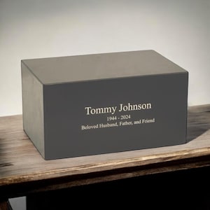 May include: A gray rectangular cremation urn with the inscription "Tommy Johnson 1944 - 2024 Beloved Husband, Father, and Friend".
