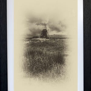 Puede incluir: Impresión enmarcada en tonos sepia de un molino de viento en un campo bajo un cielo nublado. El molino de viento es el punto focal, con hierba alta en primer plano. La impresión está en un marco negro con un passe-partout blanco.