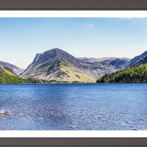 May include: A landscape photograph showing a lake with mountains in the background. The water is a deep blue, reflecting the sky. The mountains are covered in green trees and yellow-green grass. The sky is a clear blue with a few clouds.