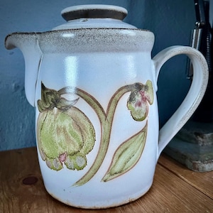 May include: A white ceramic jug with a lid and handle. The jug features green and brown floral patterns. The rim and lid have a speckled brown trim. The jug is probably used for serving drinks.