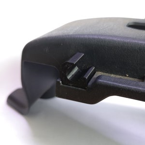 May include: A close-up of a black plastic automotive component, likely a trim piece. The part features a textured surface, a curved design, a rectangular cutout, and a small clip. The image is well-lit, showing the details of the part.