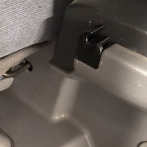 May include: Close-up of a black plastic latch or clip attached to a grey surface. The latch is designed to secure a seat or other component in place.