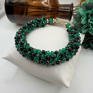May include: A bracelet made of black and green beads, woven together in a detailed pattern. The bracelet is displayed on a white cushion. The clasp is gold-toned. The background includes a brown glass bottle and green floral decorations.