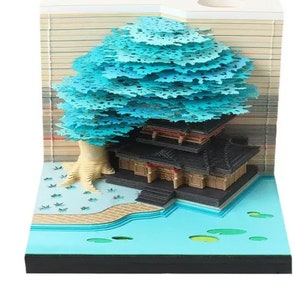 May include: A 3D papercraft notepad featuring a blue tree and a black and brown pagoda-style building. The notepad is set against a blue water background with green lily pads.