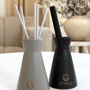 May include: Two ceramic reed diffuser bottles, one grey and one black, with white and black reeds. The grey bottle has a gold logo and the word "OPPOLI" printed on it. The black bottle also has a gold logo and the word "OPPOLI" printed on it.