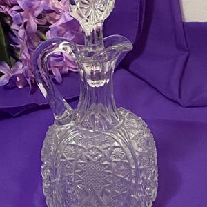 May include: A clear, cut-glass decanter with a handle and stopper. The decanter has a rounded body with a detailed starburst pattern. The stopper is a round shape with a similar starburst design. The decanter is on a purple surface.