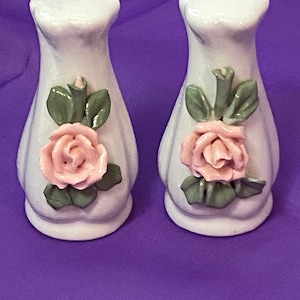 May include: A pair of white ceramic salt and pepper shakers. Each shaker features a pink rose with green leaves. The shakers have a classic design and are suitable for a dining table.