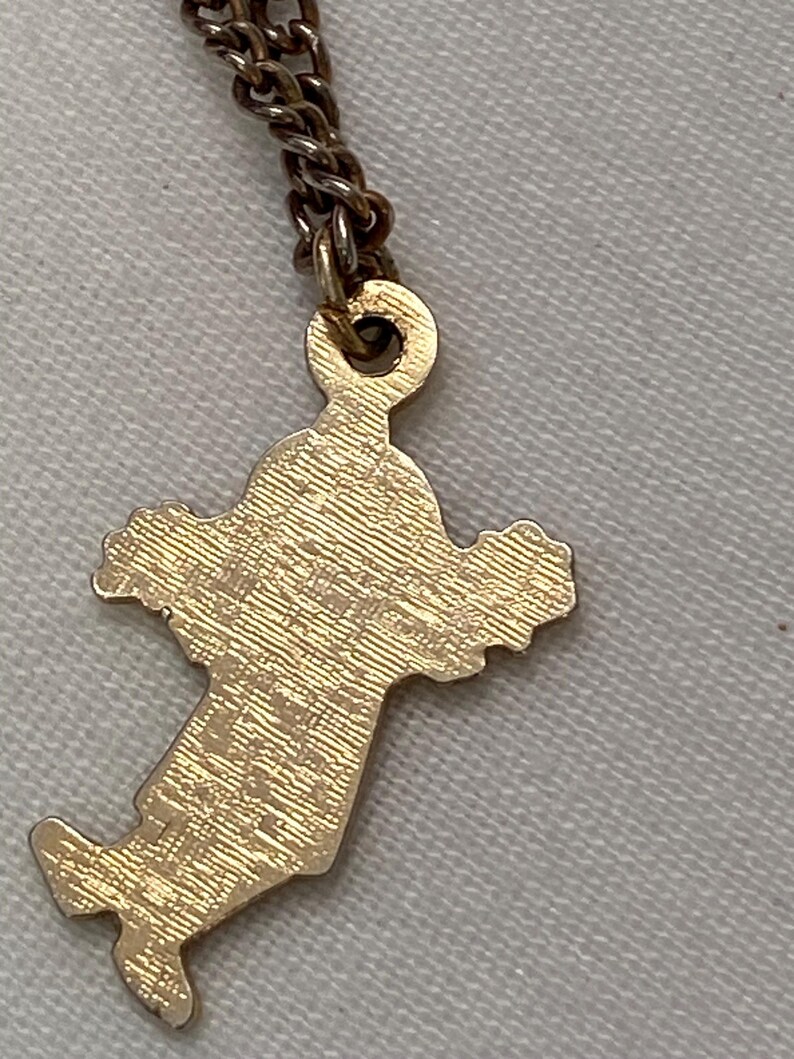 May include: A gold-colored pendant in the shape of Italy hangs from a delicate chain. The pendant has a textured surface and is attached to a small, circular loop. The chain is a darker, bronze color.