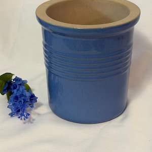 May include: A cylindrical ceramic crock with a glossy blue exterior finish and a series of horizontal ridges around the upper body. The interior is a smooth, unglazed beige color. A small cluster of artificial blue flowers sits beside the base.