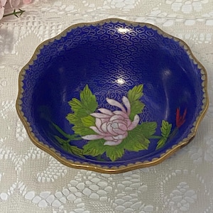 May include: A small, round, blue decorative bowl with a gold rim. The bowl features a floral design with a pink flower and green leaves. The interior has a repeating pattern. A beautiful piece for display.