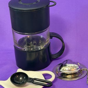 May include: A black and clear glass tea infuser mug with a handle, accompanied by a white ceramic spoon rest holding a black plastic scoop, and a silver-toned heart-shaped metal tea infuser resting on a small circular metal drip tray.