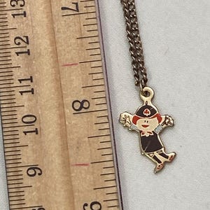 May include: A gold-toned necklace with a pendant featuring a cartoon figure in a dark uniform and red hat. The pendant is approximately 1 inch in height. The chain is a delicate, linked design.