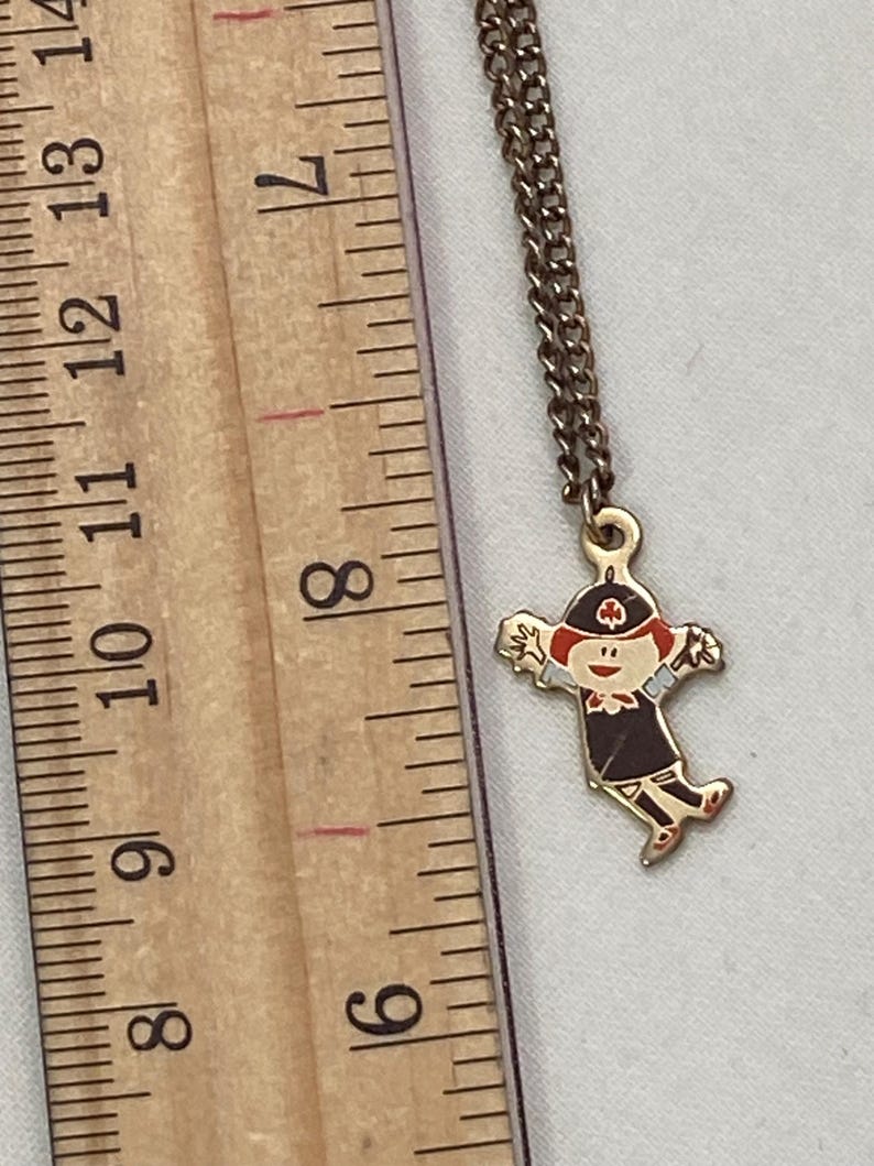 May include: A gold-toned necklace with a pendant featuring a cartoon figure in a dark uniform and red hat. The pendant is approximately 1 inch in height. The chain is a delicate, linked design.