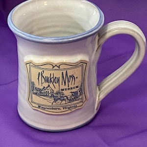 May include: A ceramic mug with a light blue rim and handle, featuring a label with the text "P. Buckley Moss Museum" and an illustration of a building and horse-drawn carriage. The mug is white with a light blue accent.