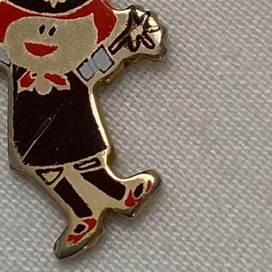 May include: A gold-toned pin in the shape of a cartoon character. The character is wearing a black uniform with red accents, a black hat, and red shoes. The pin has a smiling face and is holding a flower.