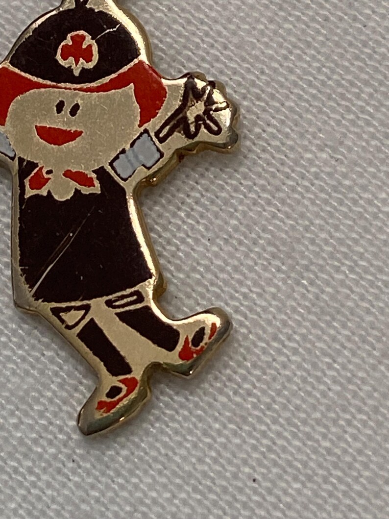 May include: A gold-toned pin in the shape of a cartoon character. The character is wearing a black uniform with red accents, a black hat, and red shoes. The pin has a smiling face and is holding a flower.