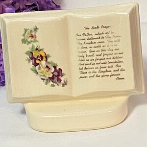 May include: Cream-colored ceramic book-shaped decorative piece. The open pages display the text of "The Lord's Prayer" in black script. Floral accents of purple, yellow, and white pansies adorn the left page. The base is curved.
