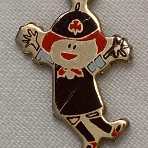 May include: A gold-toned charm in the shape of a cartoon figure. The figure has red hair, a black hat with a red cross, and a black uniform. The charm has outstretched arms and red shoes. The charm is attached to a gold-toned loop.