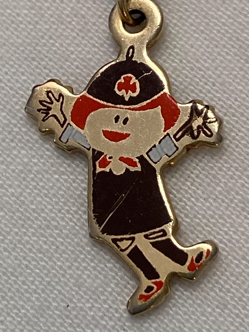 May include: A gold-toned charm in the shape of a cartoon figure. The figure has red hair, a black hat with a red cross, and a black uniform. The charm has outstretched arms and red shoes. The charm is attached to a gold-toned loop.