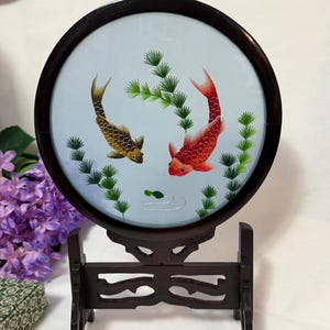 May include: A circular embroidery piece featuring a gold and a red koi fish swimming among green aquatic plants on a light blue background, encased in a dark brown wooden frame and supported by a matching decorative wooden stand.