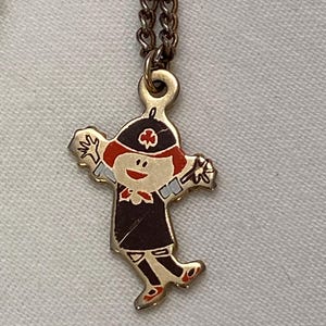 May include: A vintage gold-tone pendant necklace featuring a cartoon figure in a black dress, red hair, and a hat with a cross emblem. The figure is in a playful pose with arms outstretched. The necklace chain is a matching gold color.