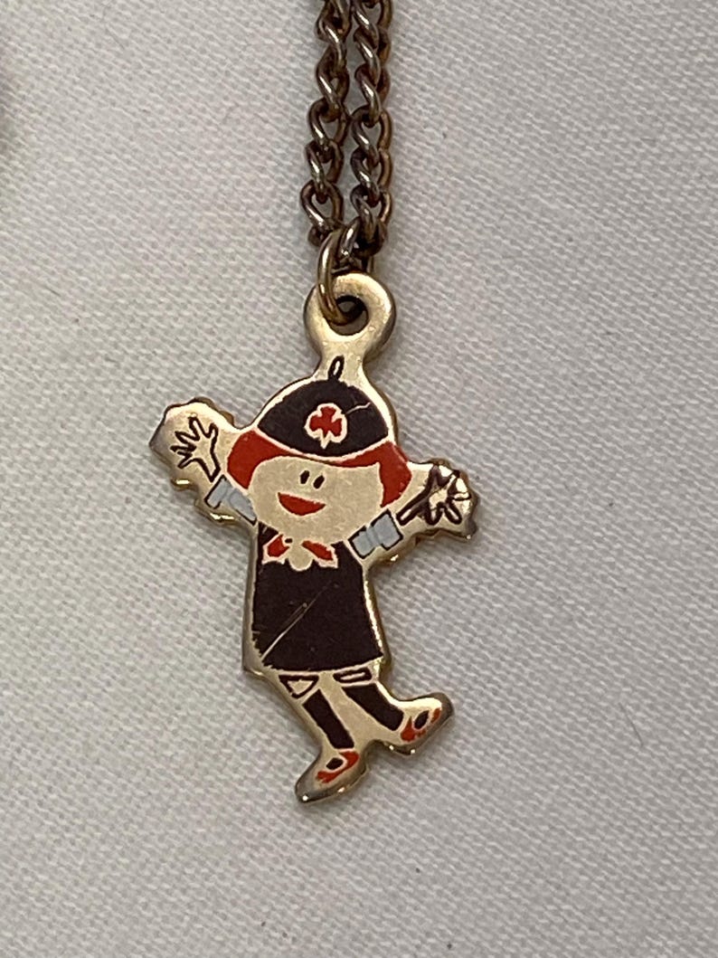 May include: A vintage gold-tone pendant necklace featuring a cartoon figure in a black dress, red hair, and a hat with a cross emblem. The figure is in a playful pose with arms outstretched. The necklace chain is a matching gold color.