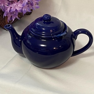 May include: A glossy, navy blue ceramic teapot with a rounded body, curved handle, and a spout. The lid has a small knob. The teapot is set against a white background, with a basket of purple flowers in the background.
