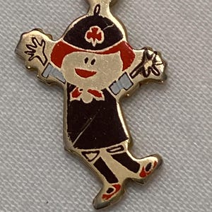 May include: A gold-colored metal charm in the shape of a cartoon figure. The figure has red hair, a black hat with a clover, and a dark brown outfit. The charm has outstretched arms and red shoes.
