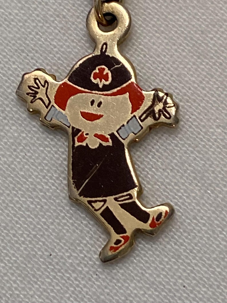May include: A gold-colored metal charm in the shape of a cartoon figure. The figure has red hair, a black hat with a clover, and a dark brown outfit. The charm has outstretched arms and red shoes.
