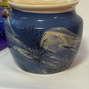 May include: A blue ceramic vase with a cream-colored rim. The vase features a landscape design with a tree and cloud-like patterns in shades of blue and cream. The vase is a decorative item.