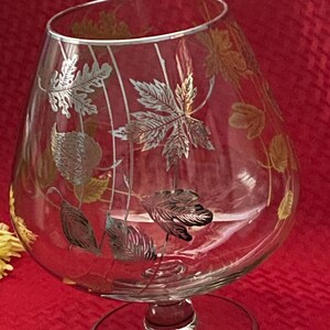 May include: A clear brandy snifter with a silver rim and a decorative leaf design in silver and gold. The glass has a wide bowl and a short stem, perfect for serving brandy or other spirits.