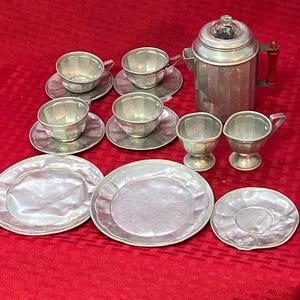 May include: A vintage silver-toned tea set, including a coffee pot with a red handle, cups, saucers, plates, and creamers. The set is arranged on a red tablecloth, showcasing a classic design.
