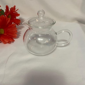 May include: A clear glass teapot with a rounded body, a curved handle, and a spout. The teapot has a clear lid with a small knob on top. Red flowers are visible in the background. The teapot is designed for brewing tea.