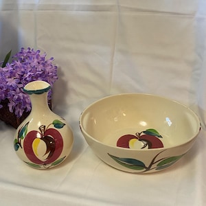 May include: Cream-colored ceramic vase and bowl set. The vase is a rounded shape with a narrow neck. The bowl is round with a wide opening. Both pieces feature a red apple design with green leaves and brown stems.