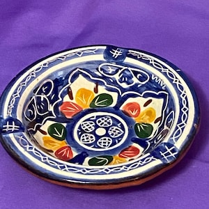 May include: A round ceramic ashtray with a vibrant, hand-painted design. The ashtray features a blue and white border, with a central pattern of flowers and leaves in red, yellow, and green. It has a small, raised edge and a brown base.