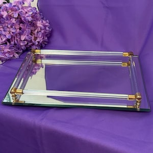 May include: A rectangular mirrored tray featuring a clear acrylic handle on each side. The handles are secured with polished gold-tone metal accents at the ends. The tray has a reflective glass surface and a clean, structured design for display.