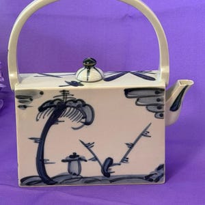 May include: A rectangular ceramic teapot with a high arched handle and a small lid. The teapot is off-white with hand-painted blue botanical illustrations, including a tree and bamboo-like stalks, and features a short spout on the right side.