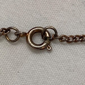 May include: Close-up of a gold-colored necklace clasp and chain. The clasp is a circular spring ring type, and the chain links are small and oval. The background is a light-colored fabric.