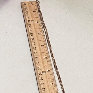 May include: A gold-toned necklace with a pendant featuring a cartoon figure. The necklace is measured with a wooden ruler, showing the chain's length. The pendant is a small, detailed design with red and black accents.